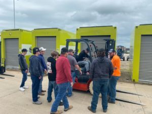 Several people are standing around a forklift learning how to use it. 