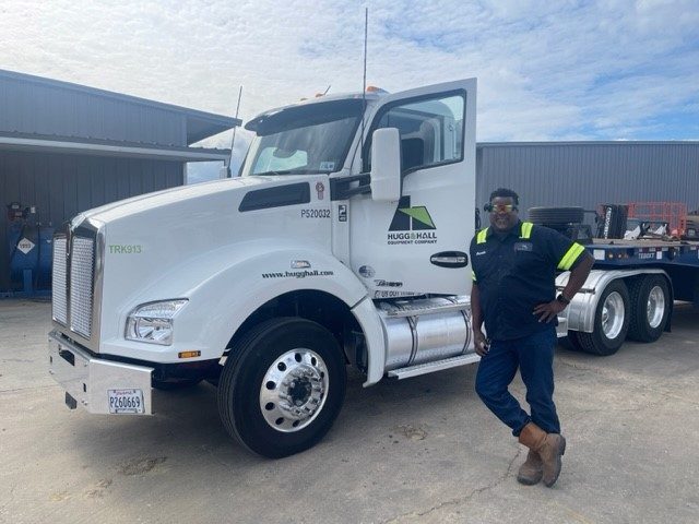 Kenneth Jones is standing next to his delivery truck, a semi cab with a trailer. He is wearing a blue uniform with yellow reflective stripes and leaning up against the open door of the semi. He is wearing sunglasses and smiling. 