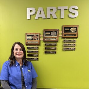 Cyndy Adams stands in front of a wall with plaques detailing Toyota Parts Professional Certifications. She is gold-level certified.