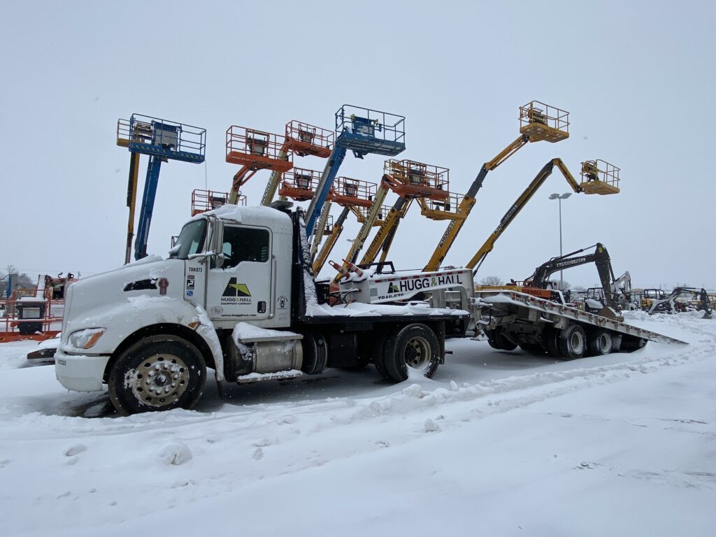 Blog post about winter construction jobs and opportunities. A Hugg & Hall delivery truck with boom lifts in the background. There is snow on the ground. 